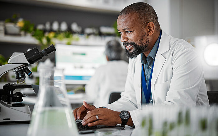 Smiling biologist using a laptop in a lab, analyzing plant growth for GMO or sustainability research.