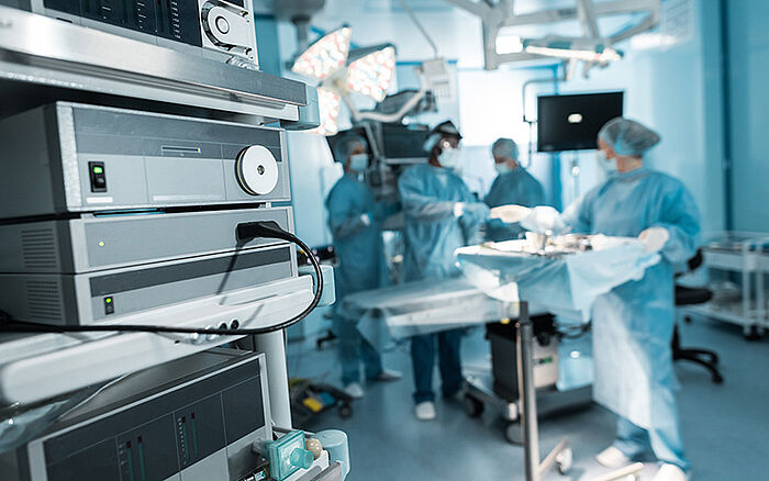 Four surgeons in scrubs preparing in a brightly lit operating room, standing around the surgical table with medical equipment in the background.